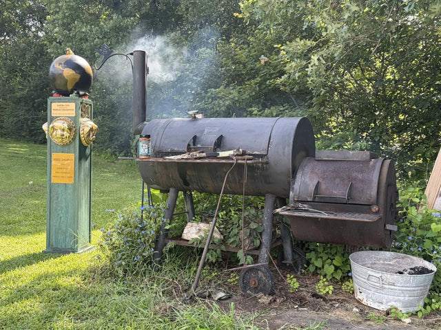 Uncle Dave's hand-welded smoker with smoke rolling, a bottle of Cox Rub on the shelf, and the 1991 World Champion trophy standing beside it